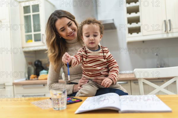 Mother holding her young toddler on her lap, together they are painting in a coloring book with watercolors and a brush, sharing a happy and educational moment in the kitchen