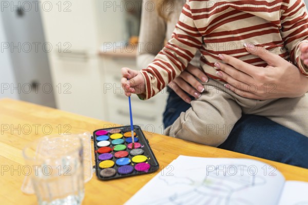 Young child held by mother, dipping a brush into a vibrant watercolor palette while coloring in an activity book, fostering creativity, bonding, and artistic development