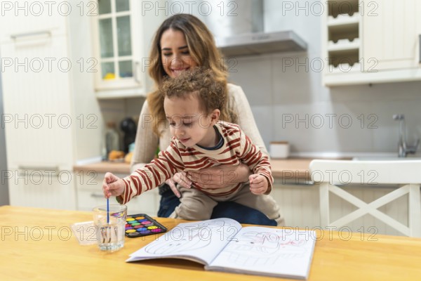 Happy mother holding son at the kitchen table as he dips a paintbrush into water, preparing to paint in a coloring book, sharing a cozy, creative moment together