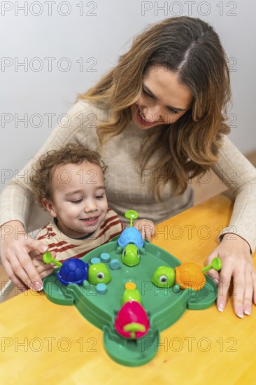 Happy mother and young son share joyful indoor play, laughing while learning with a colorful turtle themed educational board game on a wooden table, bonding and growing together