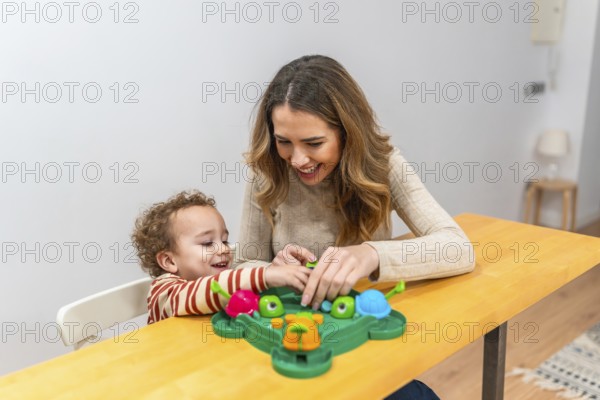 Mother and young son enjoying a playful moment, laughing and interacting while sitting at a wooden table and playing a colorful board game, sharing happiness and bonding