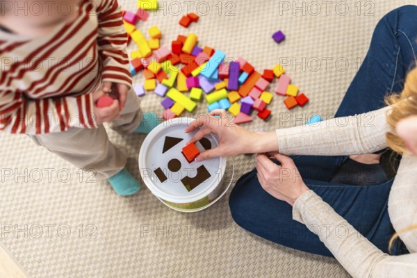 Mom assisting her toddler with a shape sorter toy and colorful blocks on a rug, fostering early childhood development, playful learning, and educational engagement at home