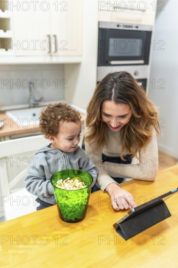 Mother and young son enjoying a family moment, sharing popcorn while engaging with a digital tablet at the wooden kitchen counter in their modern home