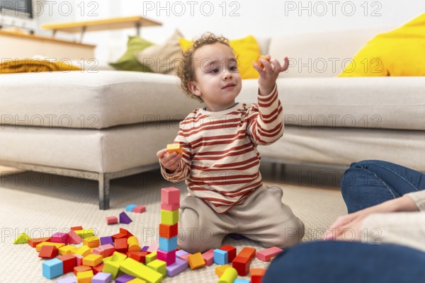 Toddler boy concentrating as he builds a tall wooden block tower on the living room carpet while an adult watches, capturing focused play, creativity and fine motor development