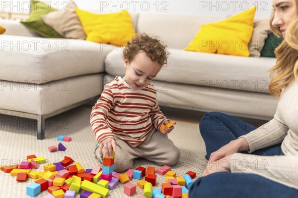 Young child with curly hair is sitting on the floor, deeply engaged in stacking colorful wooden building blocks while an adult woman watches from beside him, fostering learning and development