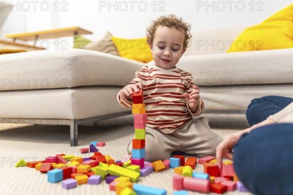 Young boy playing with colorful wooden building blocks, concentrating on constructing a tall tower on the living room floor, engaging in creative and educational play near an adult
