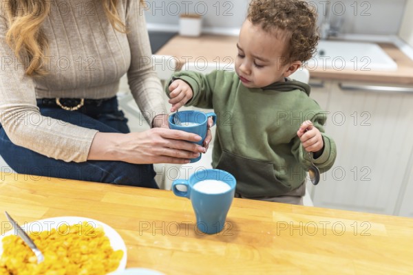 Mother guiding her toddler son stirring milk in a cup during breakfast, creating a moment around family, morning routine, and childhood in their home kitchen