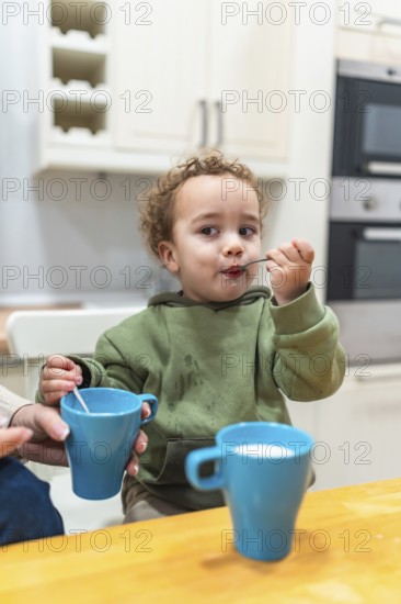 Curly haired toddler sitting at a kitchen table, independently eating yogurt from a spoon, leaving food residue on his face and green hoodie, with two blue mugs of milk beside him