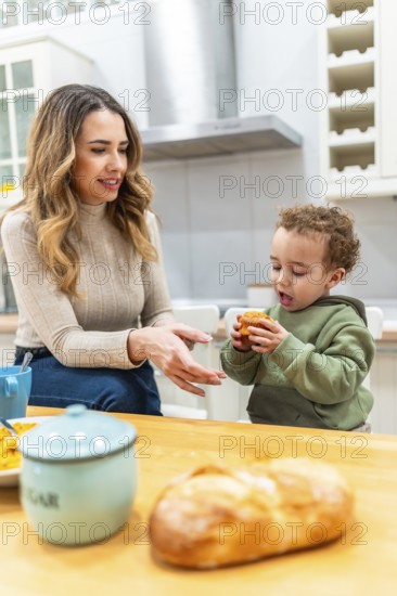 Mother looking at her happy child holding a muffin while having breakfast in a home kitchen, representing family bonding, nurturing, and early childhood development in a domestic setting