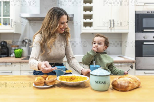 Young smiling mother feeding cereal to her adorable toddler son while sitting at the kitchen table, sharing a heartwarming family moment during morning mealtime at home
