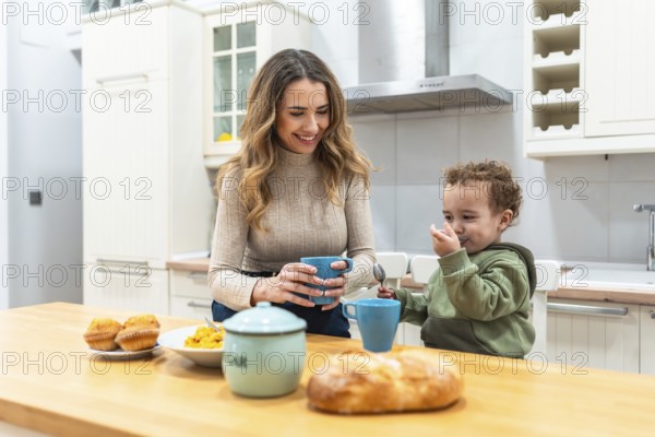 Mother and toddler share a warm morning at the kitchen counter, woman smiling with a mug while the little one eats from a bowl, cozy bonding and everyday family routine