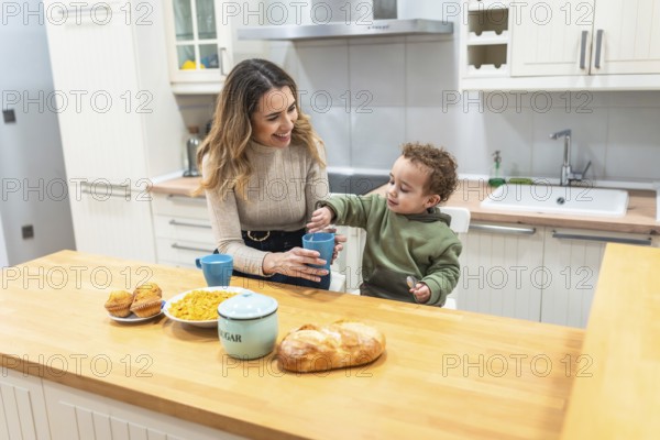 Young mother and toddler son share a cheerful breakfast at the bright kitchen island, bonding over cereal, juice and muffins in a cozy, everyday family moment