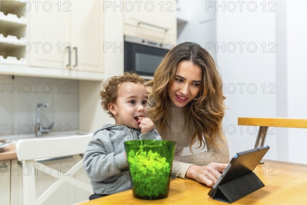 Young mother teaching and entertaining her child with a digital tablet while enjoying a snack together in a bright home kitchen, demonstrating family bonding and shared learning