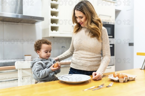 Happy mother and curious toddler boy bonding in the kitchen, preparing fresh eggs for a healthy meal while learning about cooking and nutrition together at home