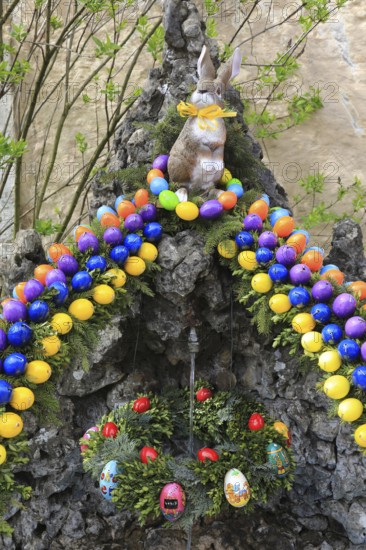 Easter fountain in Franconian Switzerland, Bamberg district, Upper Franconia, Bavaria, Germany, Easter bunny on stone arch with colorful Easter egg wreath surrounded by spring plants