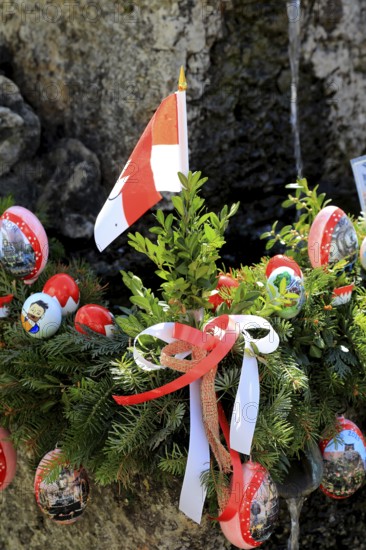 Detail of an Easter fountain in Franconian colors red and white, Franconian Switzerland, in Kleingesee, Gößweinstein market in the district of Forchheim, Upper Franconia, Bavaria, Germany, wreath with Easter eggs, spruce branches and Austrian flag as a festive decoration