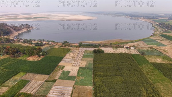 Aerial view of cultivated farmland along the riverbank, with patchwork fields stretching toward a wide river under a hazy sky