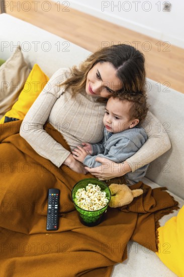 Mother holding her young son while they are relaxing on a sofa under a cozy blanket, watching a film and sharing a bowl of popcorn and creating special memories together