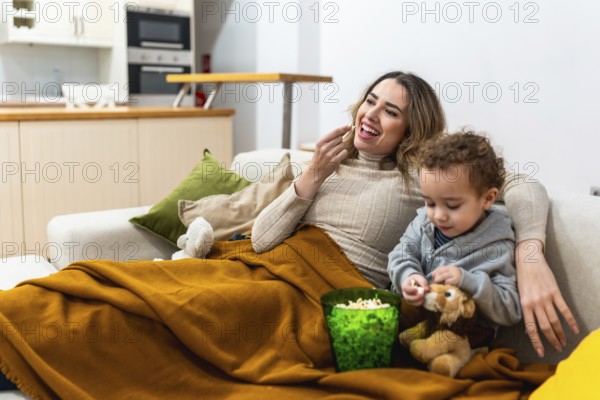 Mother and young son relaxing on a sofa under a blanket, eating popcorn and watching a movie, sharing a happy and bonding moment together in their cozy living room