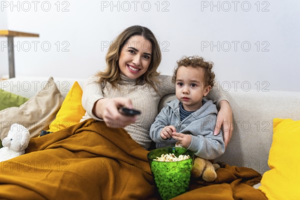 Mother and child relaxing on a cozy sofa covered by a blanket, enjoying popcorn and bonding together in their living room, using a remote control for entertainment