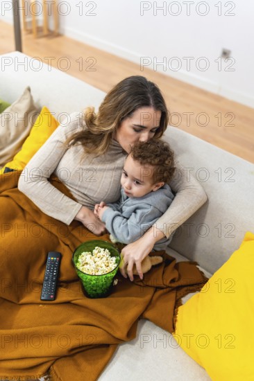 Mother and baby enjoying a bonding movie night at home, sitting comfortably on the sofa under a soft blanket while sharing popcorn and watching television