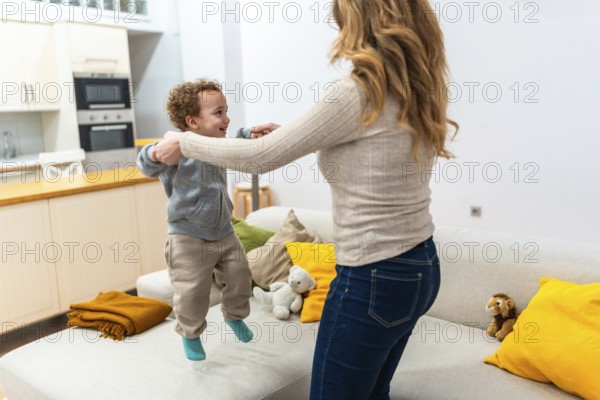 Happy mother holding her child's hands while they are jumping together on the sofa, enjoying a playful activity and bonding in the comfort of their home