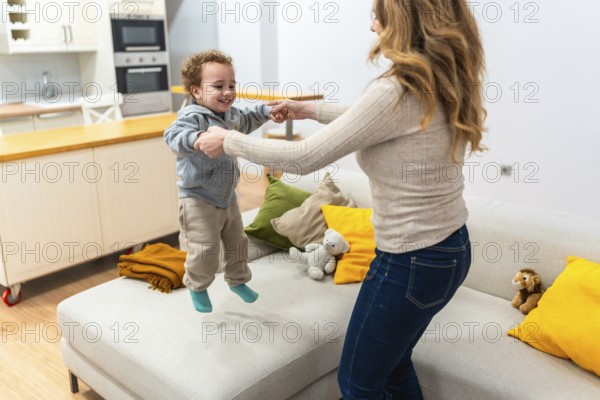 Mother and son holding hands as he joyfully jumps on the living room floor, sharing a bright, playful bonding moment of carefree childhood and warm home happiness