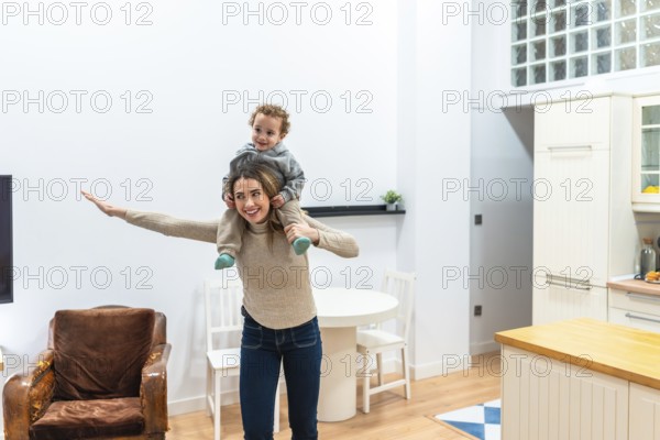 Happy mother gives toddler son a piggyback airplane ride in a bright modern apartment, sharing playful bonding, laughter and everyday joy in a cozy home setting