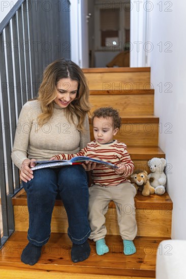 Happy mother and toddler boy enjoying reading a colorful storybook together while sitting on wooden stairs, sharing a tender moment of learning and family bonding indoors