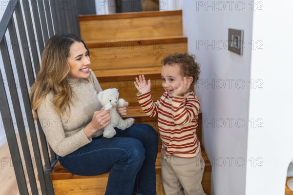 Mother and toddler sitting happily on wooden stairs, engaging in playful interaction with a small teddy bear, depicting family bonding, childhood joy, and parenting moments indoors