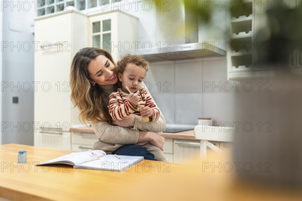 Caring mother smiling and embracing her young child while sitting at a kitchen table, bonding over a creative drawing activity with colored pencils and a sketchbook