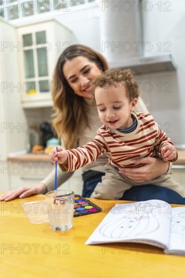 Happy mother holding her young son who is attentively painting watercolors in a coloring book, sharing a moment of family bonding and early childhood education in a domestic kitchen setting