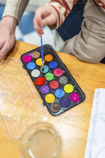 Child engaging in creative activity, dipping a small paintbrush into a colorful watercolor paint palette, with an adult hand nearby, promoting art education and hands on learning
