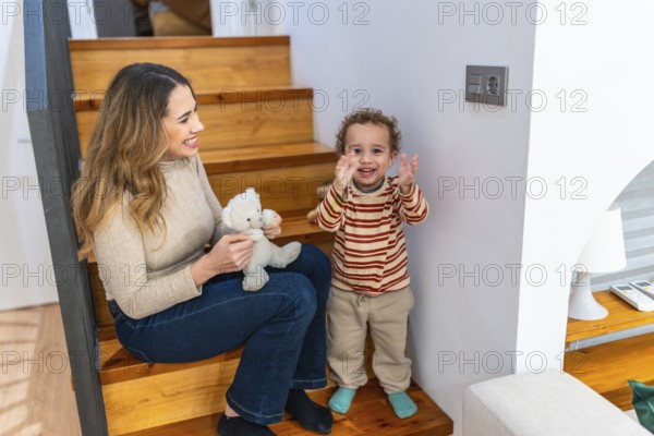 Mother and young son sit on wooden stairs at home, playing with a teddy bear and sharing a warm, joyful bonding moment of care, laughter and connection
