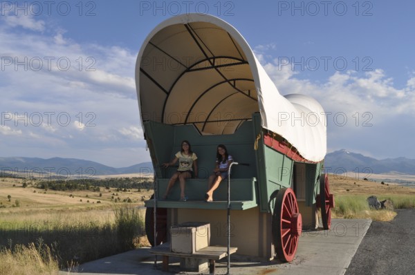 Two people sitting in a huge covered wagon, Conestoga wagon, with mountains in the background, Prairie City, Oregon, USA