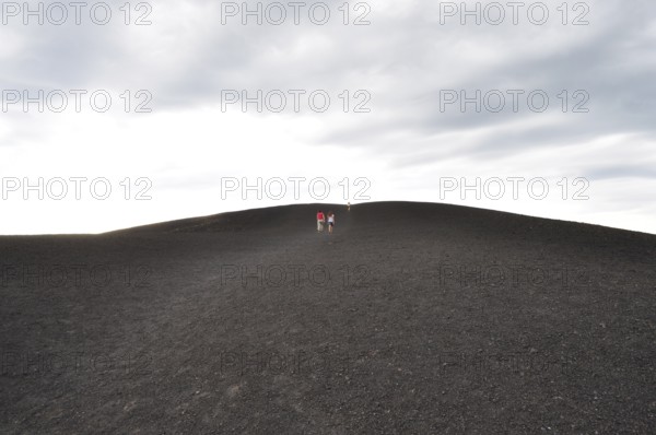 Wide landscape with people walking up a lava hill under cloudy sky, Craters of the Moon National Monument, Idaho, USA
