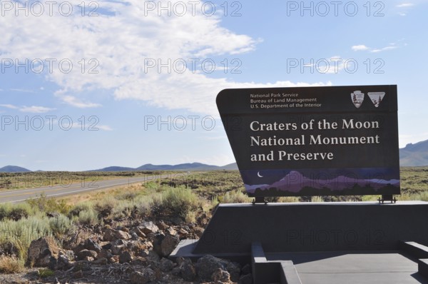 Craters of the Moon National Monument sign next to a road with wide landscape and blue sky, Craters of the Moon National Monument, Idaho, USA