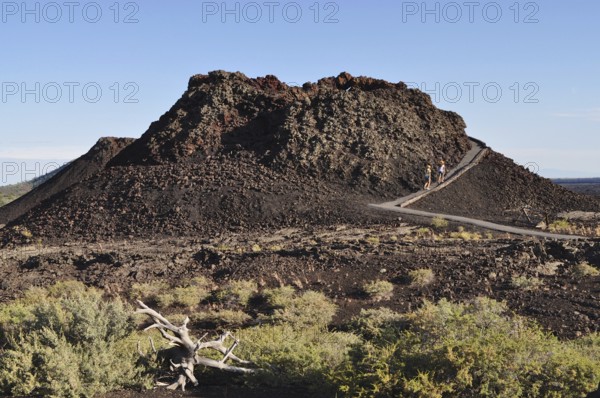 Hikers climb a volcanic mound of lava rock, sparse vegetation along the way, Craters of the Moon National Monument, Idaho, USA