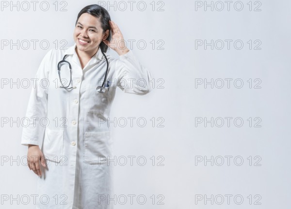 Asian doctor with hand on ear listening to a rumor isolated. Smiling female doctor hearing a rumor, looking at the camera