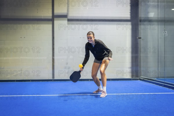 Woman athlete smiling, holding a paddle and yellow ball on a vibrant blue pickleball court, engaging in recreational sport indoors for fitness and active leisure