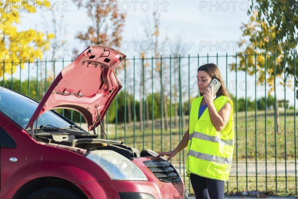 A woman wearing a reflective vest stands beside her car with an open hood, making a phone call after a breakdown The scene is set against a backdrop of trees and a fence