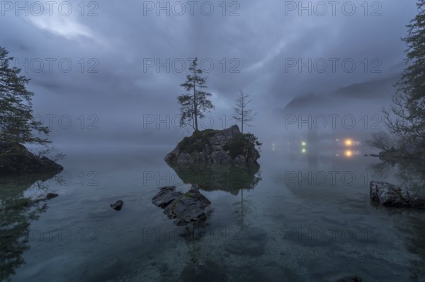 A mystical dawn view of Hintersee Lake in the Bavarian Alps, shrouded in fog with serene, still waters reflecting the cloudy sky Soft lights twinkle in the background