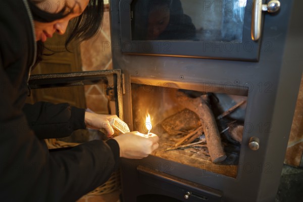 An Asian woman is seen lighting a wood burning stove, carefully holding a match to the wood. She is dressed warmly and is focused on her task, looking down towards the fire