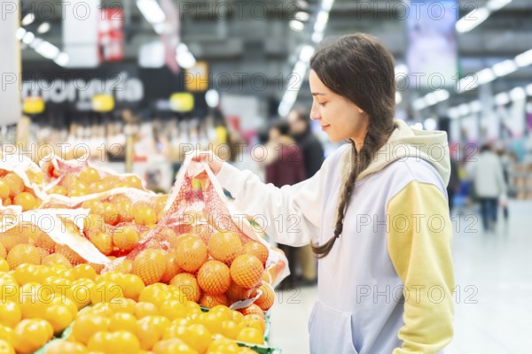 A woman is seen selecting oranges from a fresh produce section in a bustling supermarket, examining a bag of fruits carefully