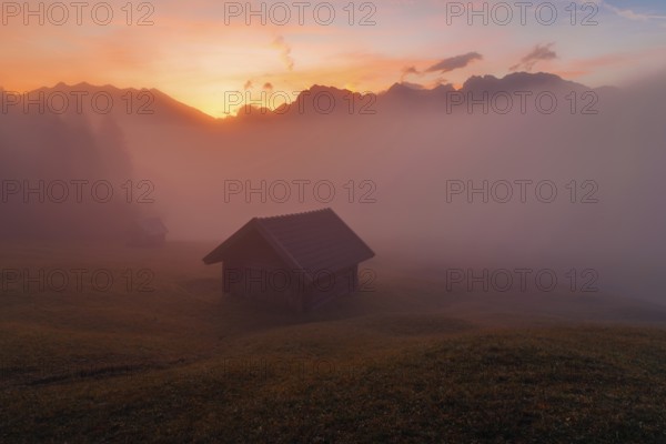A serene sunrise at Geroldsee in the Bavarian Alps Mist gently envelops wooden huts and grassy fields, creating a tranquil and picturesque landscape