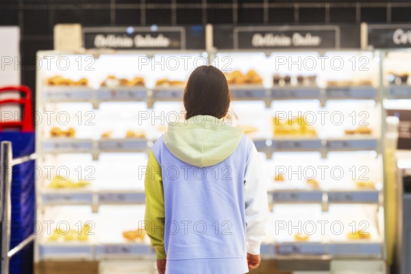 Back view of an unrecognizable woman standing in front of the bakery section in a supermarket, contemplating which product to choose