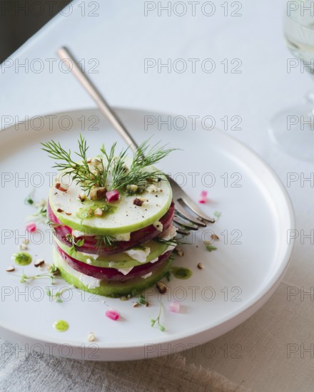 Top view of a delicately assembled gourmet salad, featuring layers of cucumber, radish, and feta cheese, garnished with dill, chopped nuts, and edible flowers on a white plate
