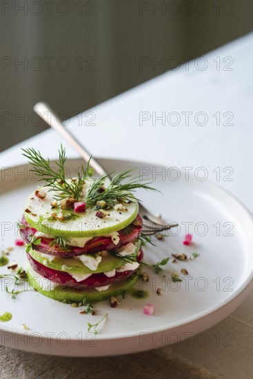 From above view of a gourmet salad featuring layers of green cucumber, red beetroot, and white cheese on a round white plate. Garnished with fresh dill, spices, and a sprinkle of crushed nuts
