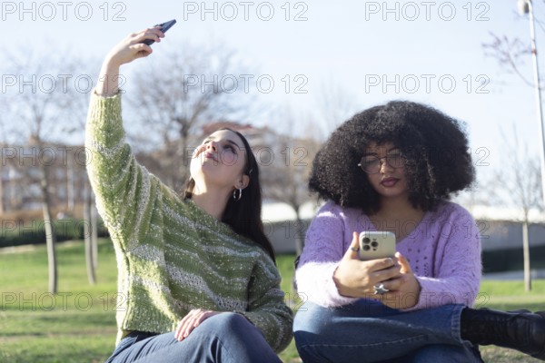 Two friends in the park, one takes a selfie while the other looks at her phone. They are enjoying a sunny day outdoors, wearing casual sweaters