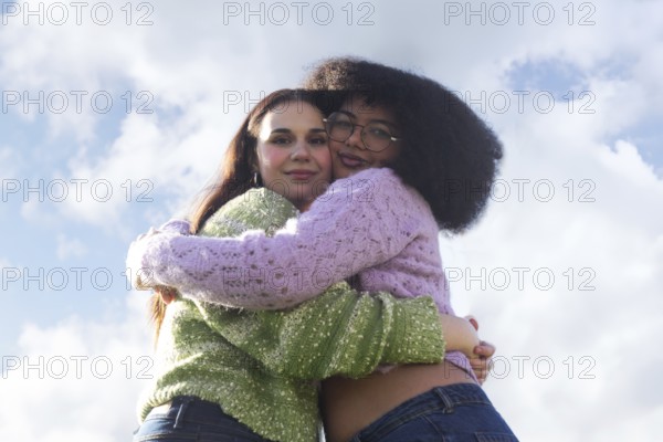 Two friends embrace warmly against a clear blue sky with fluffy clouds, wearing colorful knit sweaters. The scene radiates warmth, joy, and connection in a natural setting